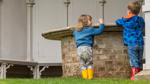 Two children in rainwear standing at brick and stone circular table nside the white painted Gothic pavilion at Uppark, Sussex